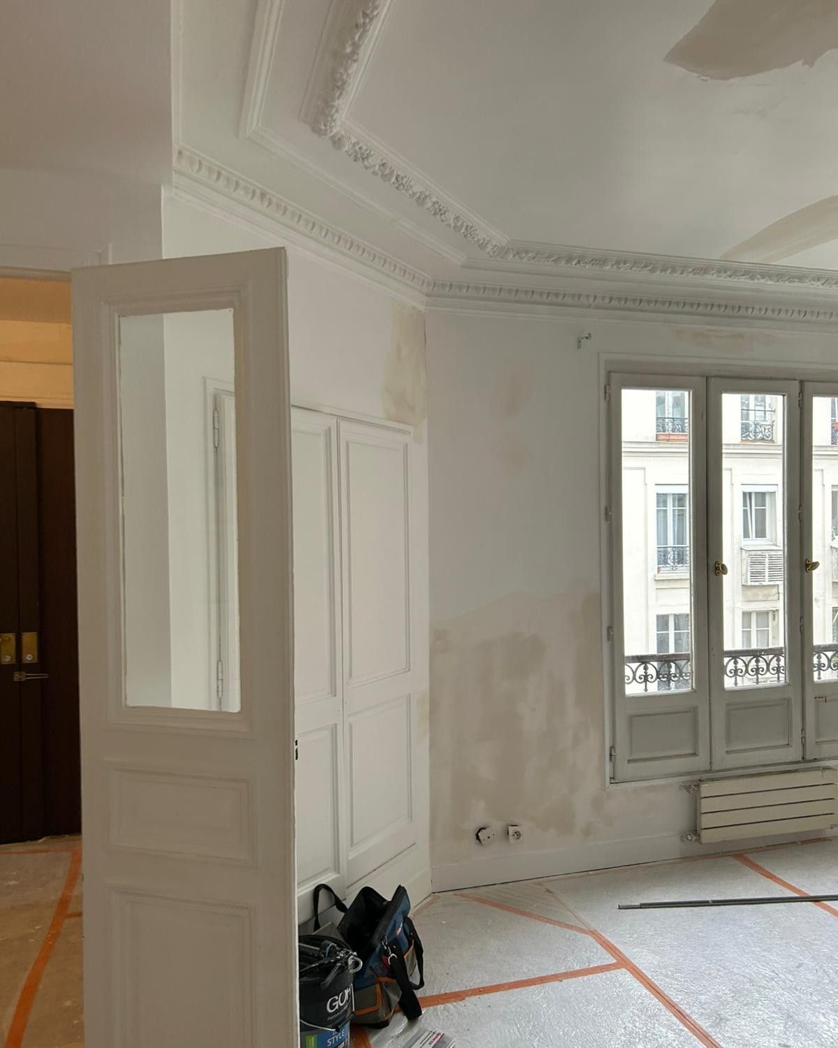 Pièce d’un appartement parisien en rénovation : corniches moulurées au plafond, porte blanche ouverte, murs repris à l’enduit et sol protégé par un film orange