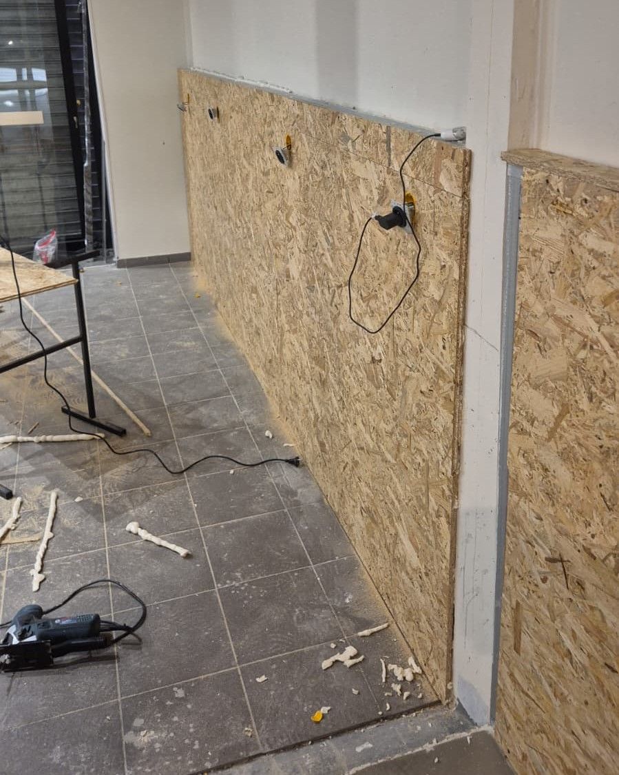 Mur blanc garni de panneaux de bois brut le long du mur, sur un sol en carrelage gris, dans une boutique et un atelier en rénovation