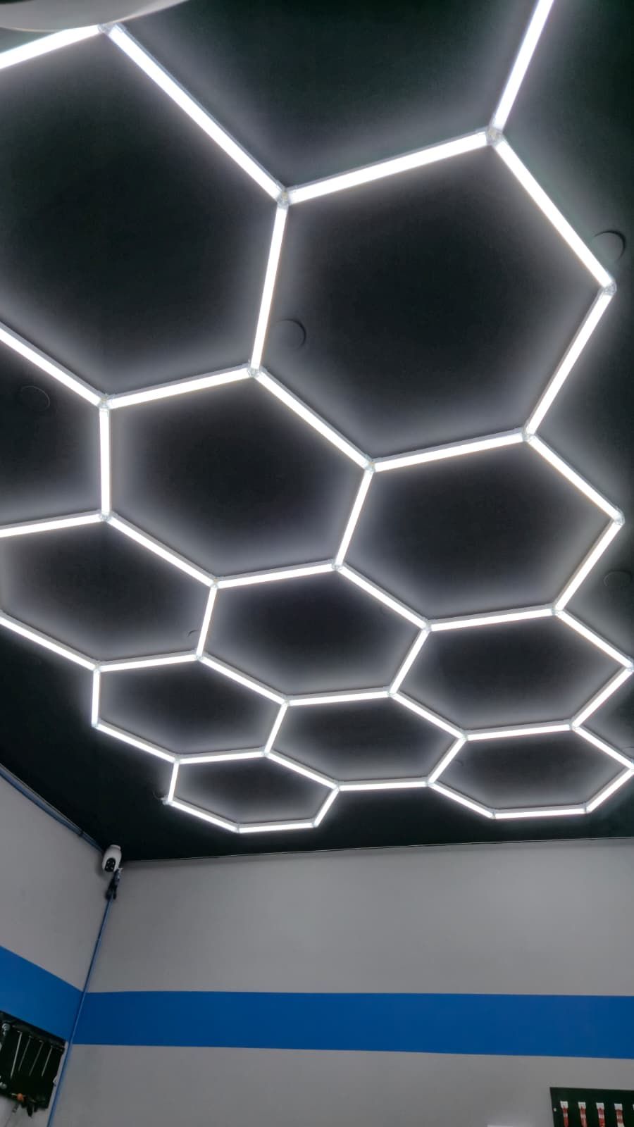 Plafonnier lumineux en forme d’alvéoles hexagonales sur un plafond noir, au-dessus d’un mur clair barré d’une bande bleue