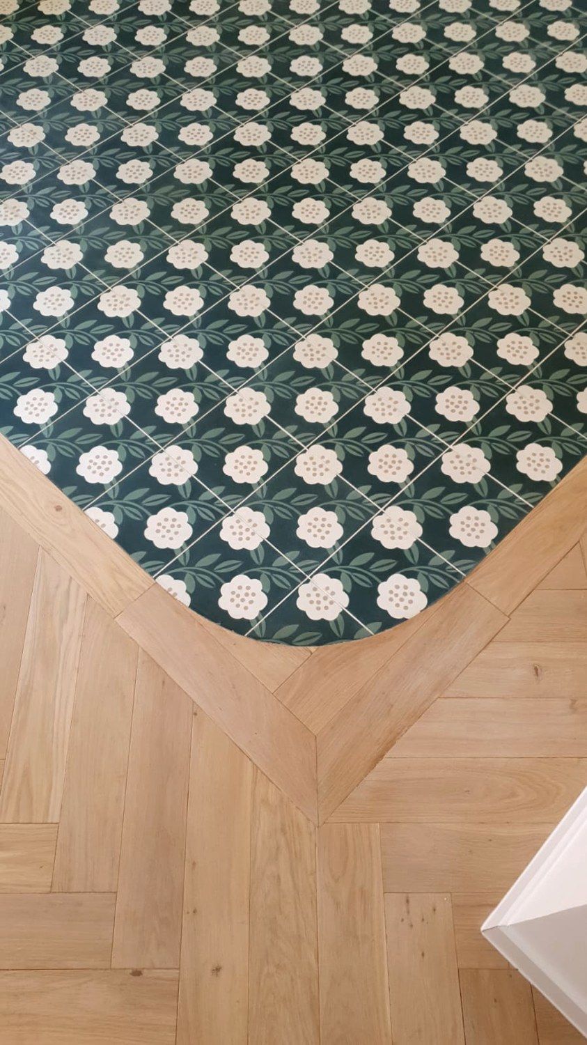 Raccord entre un parquet en chevrons et un sol en carreaux verts à motif de fleurs, avec une bordure en bois qui suit le contour
