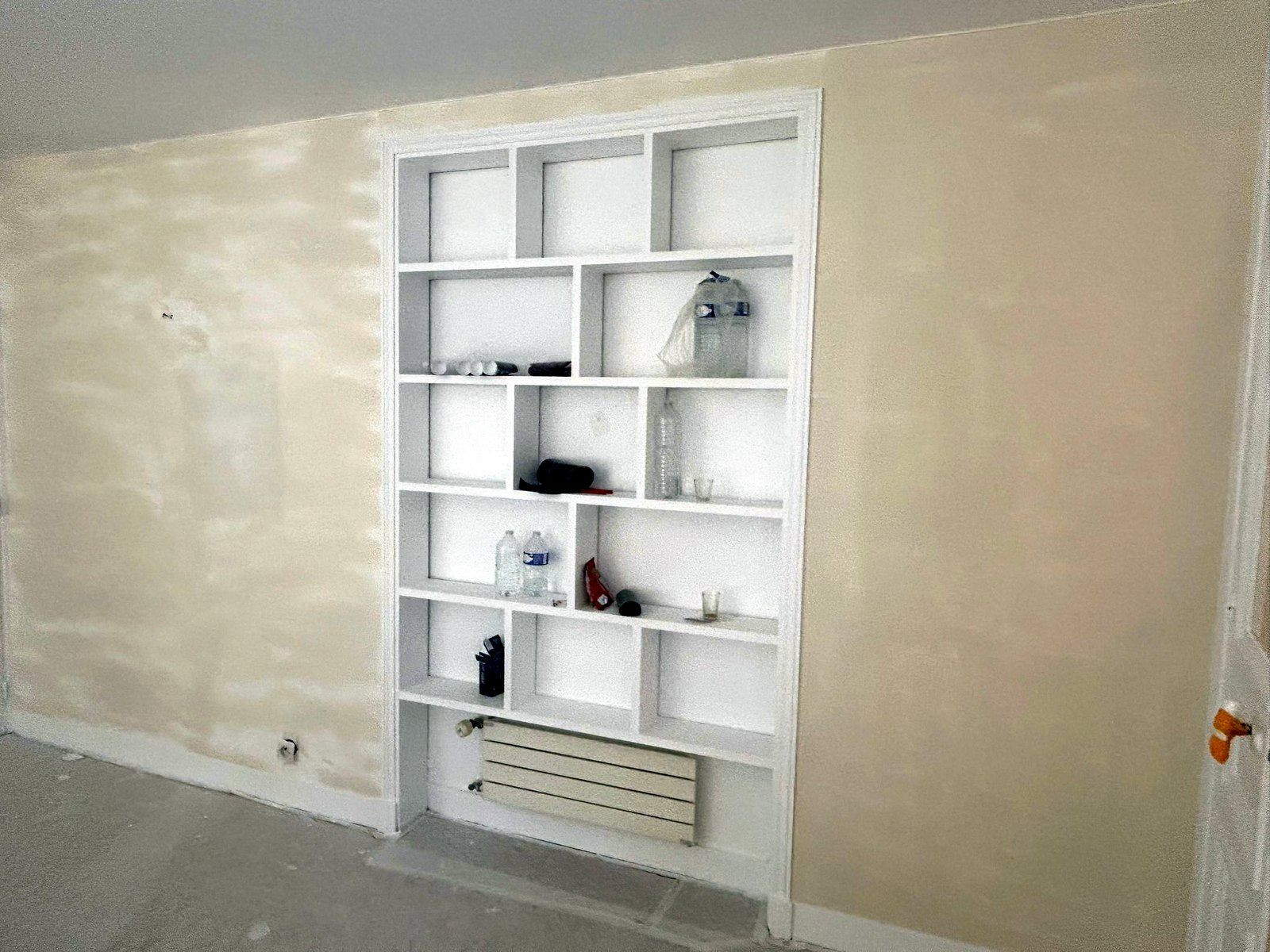 Bibliothèque encastrée blanche dans un mur repris à l’enduit, radiateur horizontal en partie basse