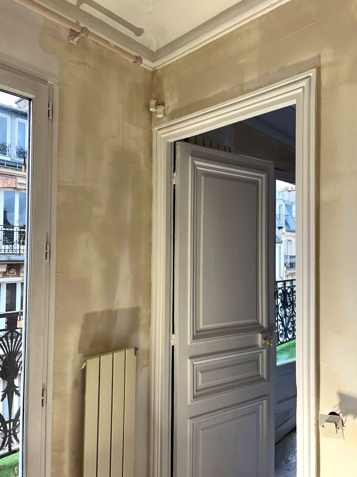 Angle d’une pièce avec une porte ouverte à moulures et une porte-fenêtre : murs repris à l’enduit, radiateur vertical