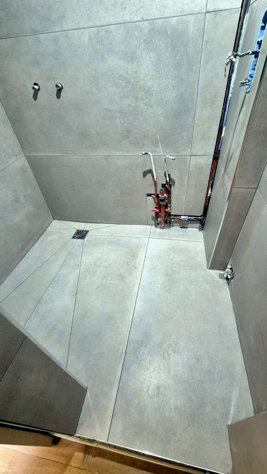 Douche carrelée en grands carreaux gris : sol aux découpes en pente vers la bonde, murs carrelés, arrivées d’eau en cuivre en attente