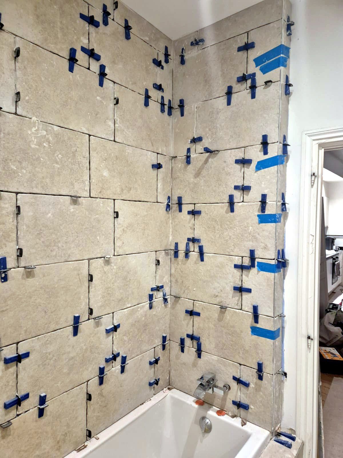 Murs d’une salle de bain en cours de carrelage : grands carreaux beiges à bords irréguliers calés par des croisillons bleus au-dessus d’une baignoire