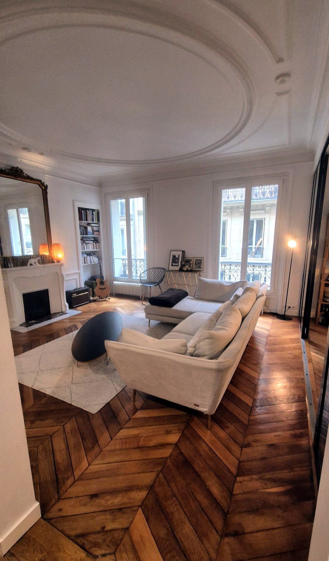 Salon d’un appartement parisien : rosace au plafond, cheminée blanche, bibliothèque encastrée, hautes fenêtres et parquet en chevrons