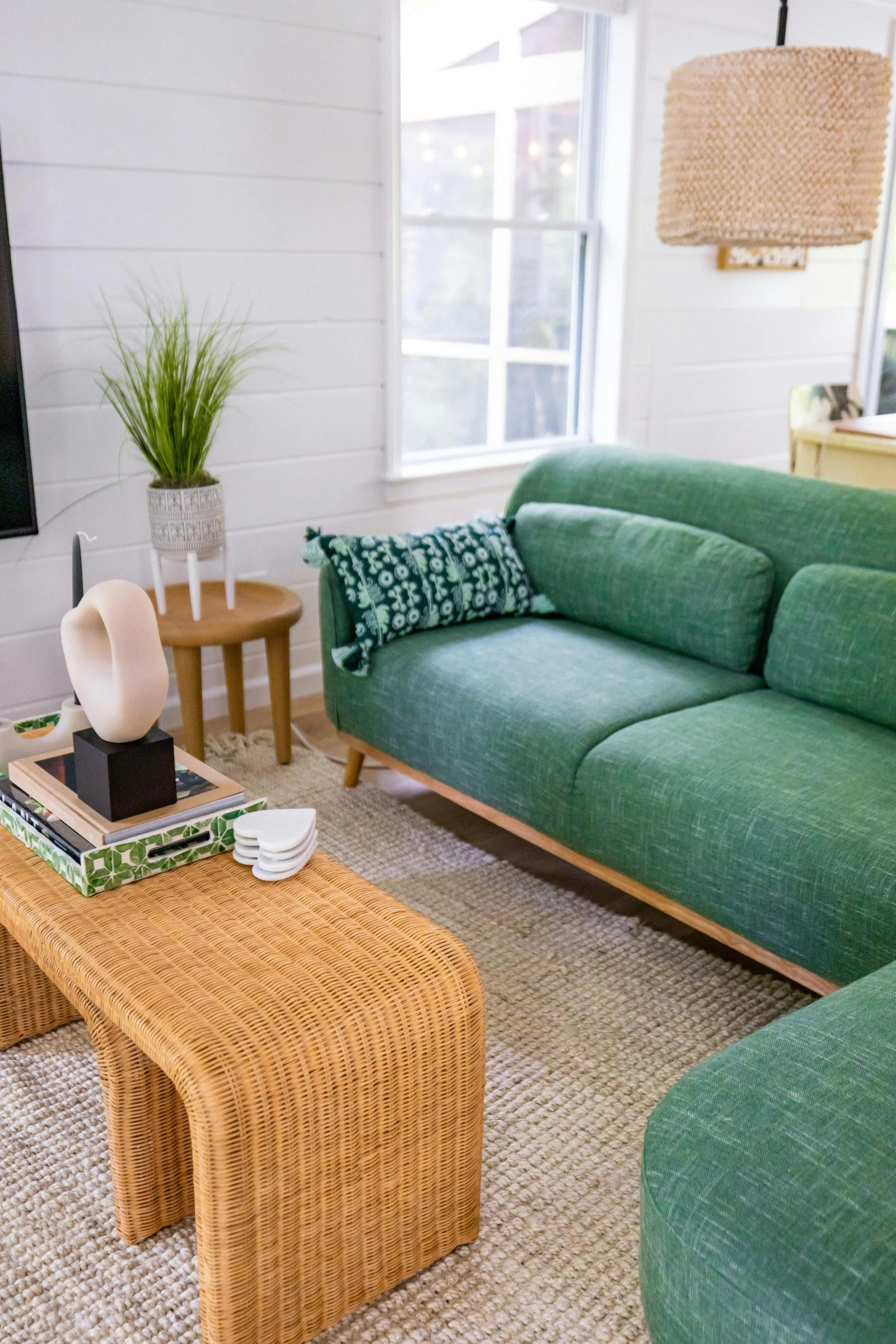 Salon aux murs blancs avec un canapé vert, une table basse en rotin, un tapis en jute et une suspension tressée