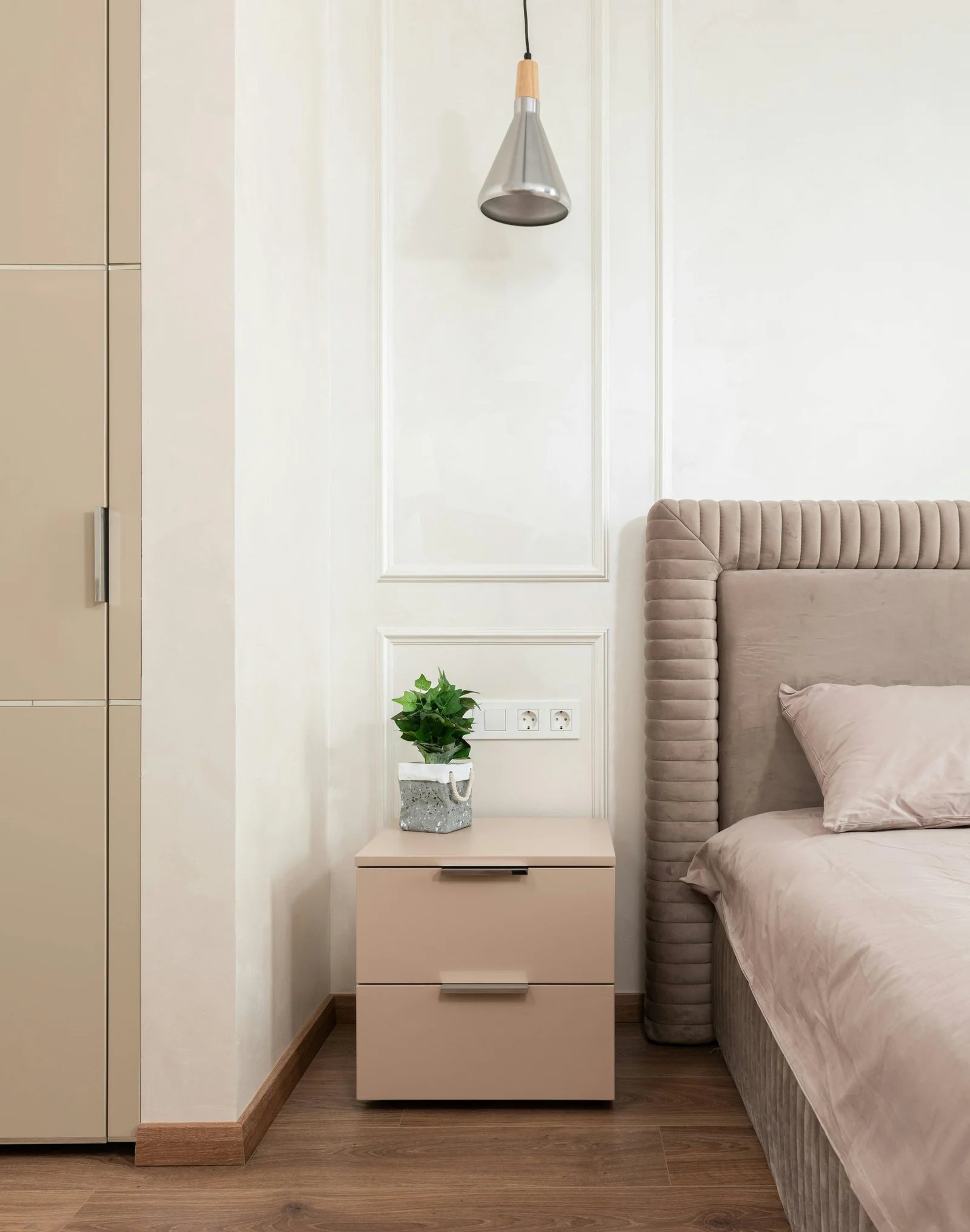 Mur clair orné de moulures, une table de chevet beige avec une petite plante et une tête de lit capitonnée