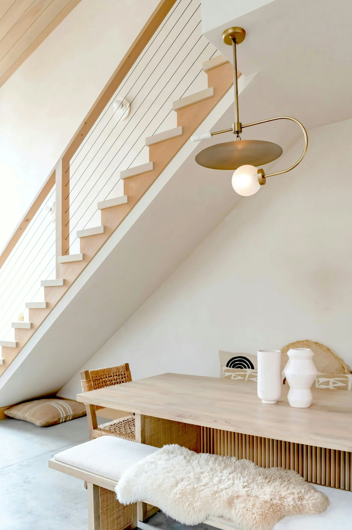 Escalier en bois clair avec garde-corps à câbles au-dessus d’une table en bois et d’une suspension en laiton