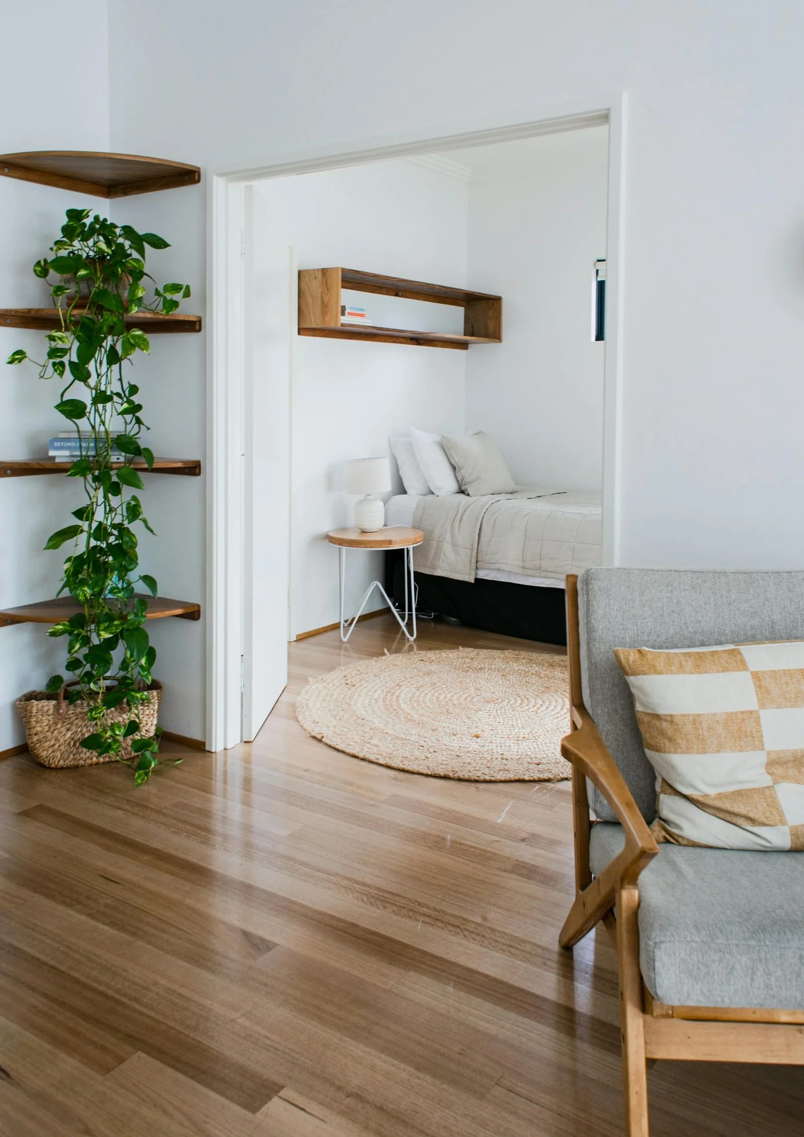 Étagères d’angle en bois garnies de plantes retombantes, avec une chambre visible par l’embrasure d’une porte et un parquet clair