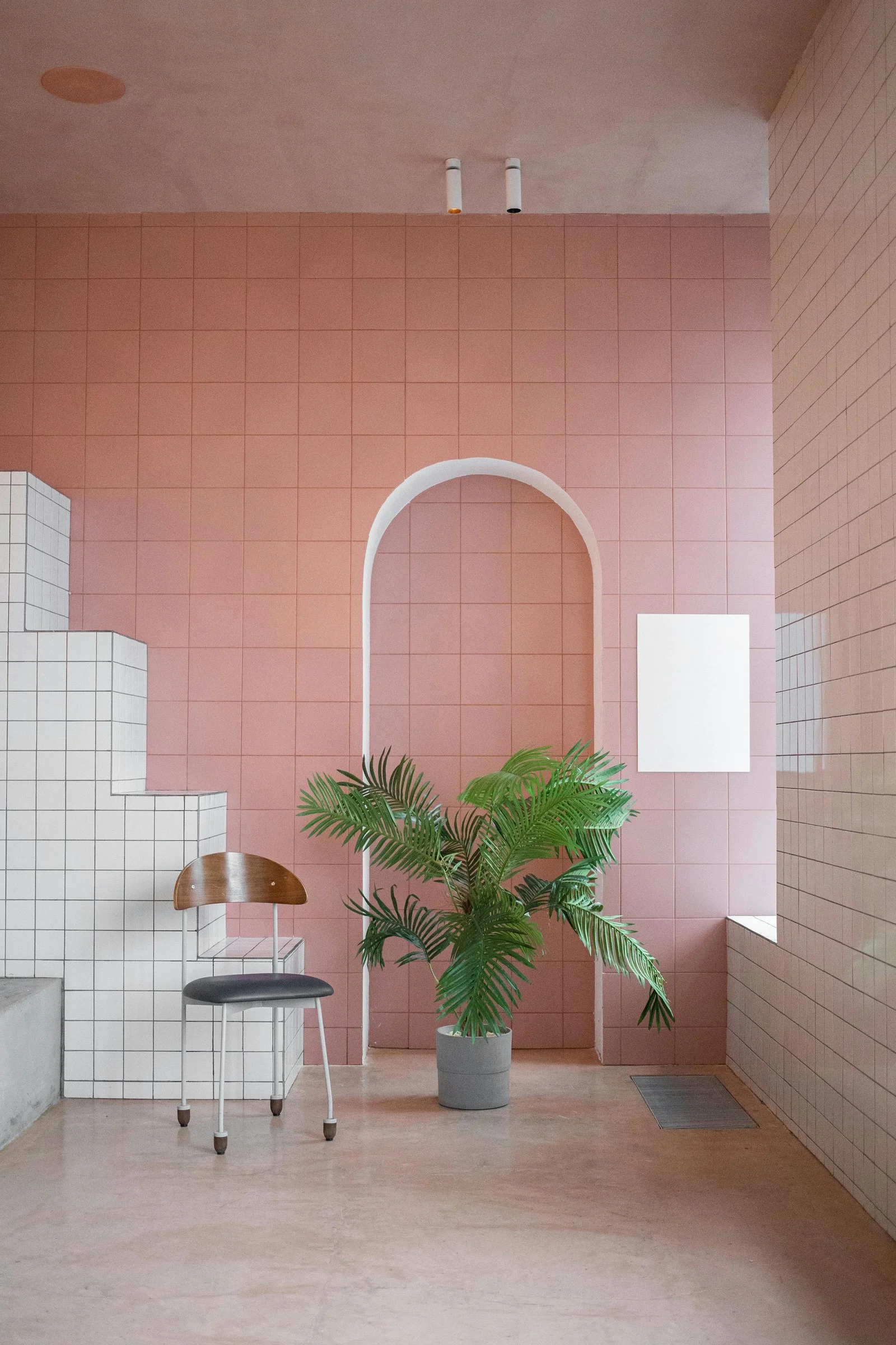 Mur rose poudré en carreaux avec une niche en arche blanche, une chaise en métal, une plante verte et des marches carrelées de blanc