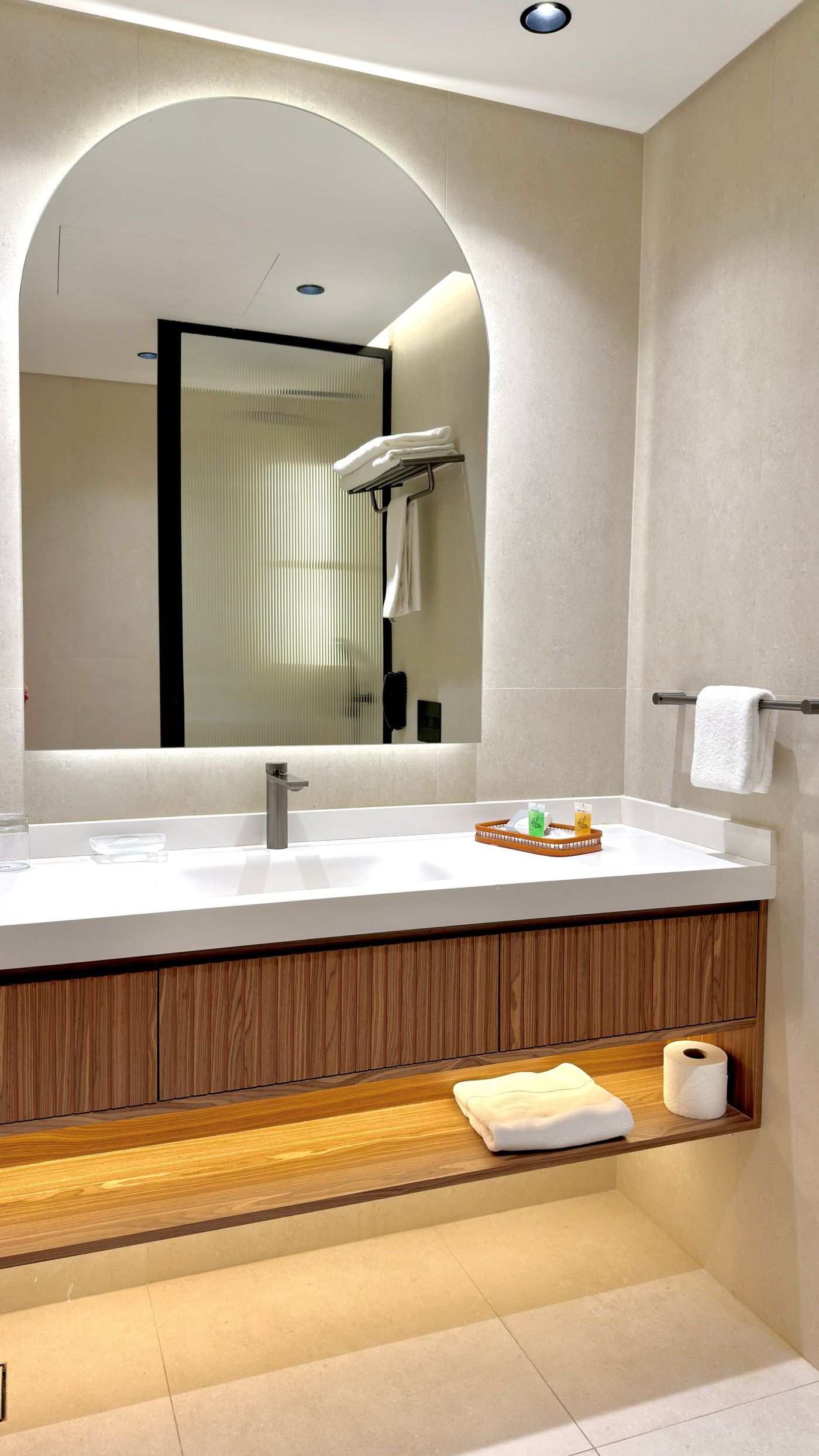 Salle de bain avec vasque suspendue sur meuble en bois, grand miroir arrondi rétroéclairé et grands carreaux clairs