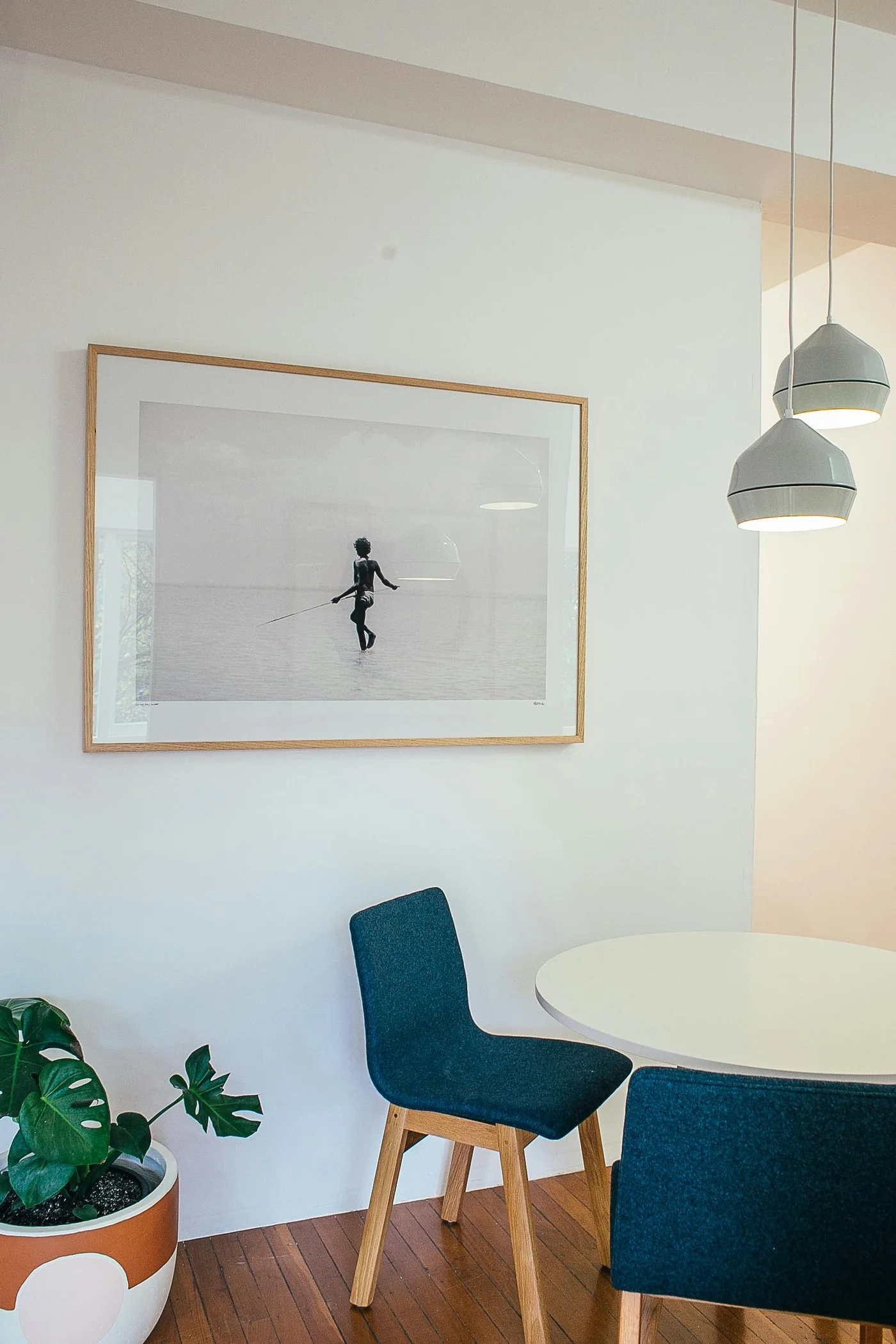 Salle à manger aux murs blancs avec un grand cadre, des chaises bleu profond, une table ronde et un parquet