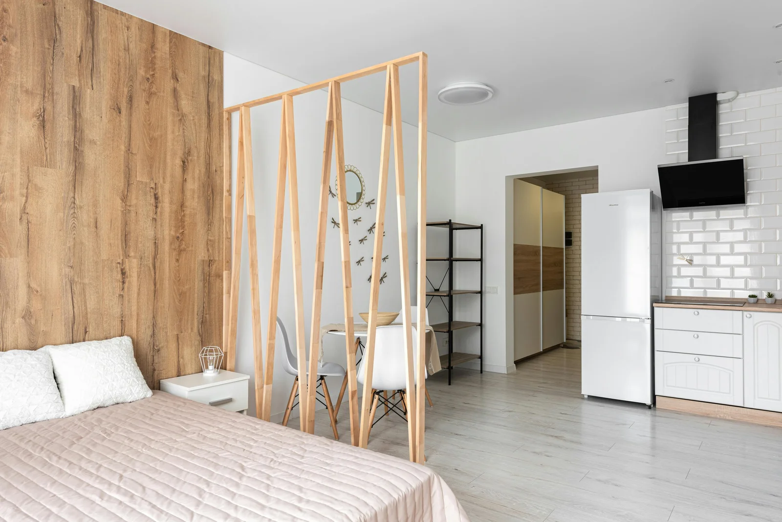Studio avec un mur en bois derrière le lit, une cloison à lattes verticales en bois et une cuisine aux carreaux métro au fond